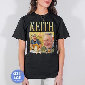 May include: Black t-shirt featuring a graphic of a man with the name "Keith" in gold lettering. The graphic includes a portrait of the man and the words "Seymour Jones". The shirt is worn by a person.
