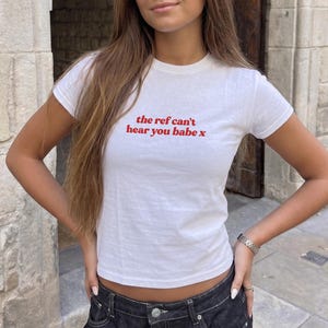 May include: A white short-sleeved t-shirt featuring the red text "the ref can't hear you babe x" printed on the chest. The shirt has a crew neckline and a fitted silhouette, paired with dark denim jeans that have a visible metal button closure.