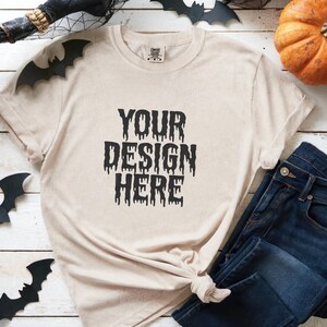 May include: A beige t-shirt with the text "YOUR DESIGN HERE" in black, paired with dark blue jeans. Halloween decorations, including bats and a pumpkin, are arranged around the clothing on a white wooden surface.