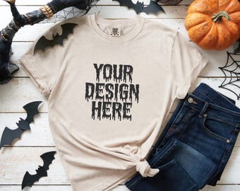 May include: A beige t-shirt with the text "YOUR DESIGN HERE" in black, paired with dark blue jeans. Halloween decorations, including bats and a pumpkin, are arranged around the clothing on a white wooden surface.