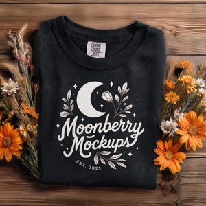 May include: A black sweatshirt with a white crescent moon and floral design, with the text "Moonberry Mockups" and "EST. 2025". The sweatshirt is surrounded by dried flowers and orange daisies on a wooden surface.