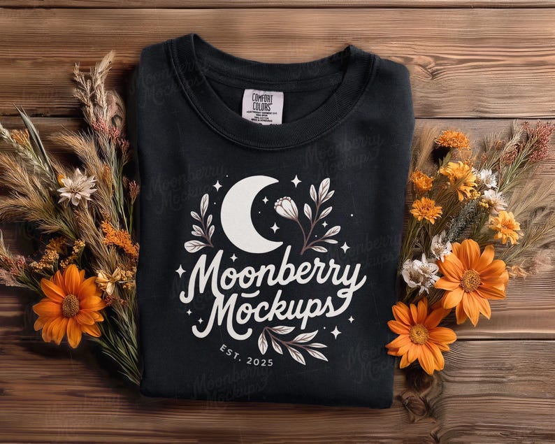 May include: A black sweatshirt with a white crescent moon and floral design, with the text "Moonberry Mockups" and "EST. 2025". The sweatshirt is surrounded by dried flowers and orange daisies on a wooden surface.