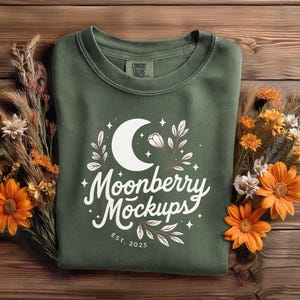 May include: An olive green sweatshirt with a white graphic featuring a crescent moon, stars, and floral elements. The text "Moonberry Mockups" is displayed in a script font, with "EST. 2025" below. The sweatshirt is surrounded by orange and yellow flowers and dried grasses.