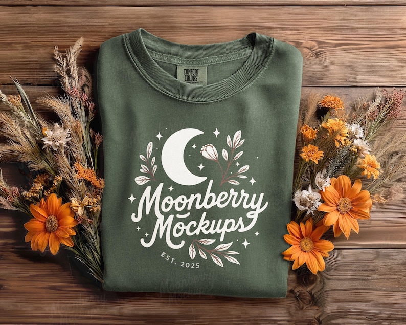 May include: An olive green sweatshirt with a white graphic featuring a crescent moon, stars, and floral elements. The text "Moonberry Mockups" is displayed in a script font, with "EST. 2025" below. The sweatshirt is surrounded by orange and yellow flowers and dried grasses.
