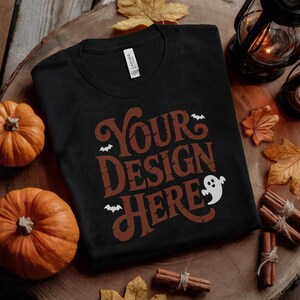 May include: Black t-shirt with the text "Your Design Here" in a distressed brown font, featuring bats and a ghost. The shirt is surrounded by pumpkins, fall leaves, and lanterns, creating a Halloween theme.