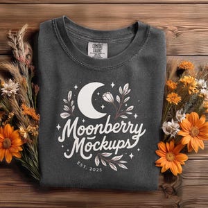 May include: A dark gray crew neck sweatshirt with the text "Moonberry Mockups" and a crescent moon design. The sweatshirt is surrounded by orange flowers and dried plants on a wooden surface.