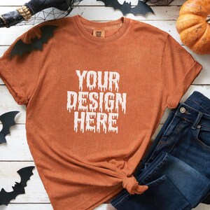 May include: An orange t-shirt with the text "YOUR DESIGN HERE" in white, paired with dark blue jeans. Halloween-themed decorations, including bats and a pumpkin, are arranged around the clothing.