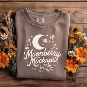 May include: A brown sweatshirt with a white crescent moon and floral design, with the text "Moonberry Mockups" and "EST. 2025". The sweatshirt is surrounded by orange flowers and dried plants on a wooden surface.