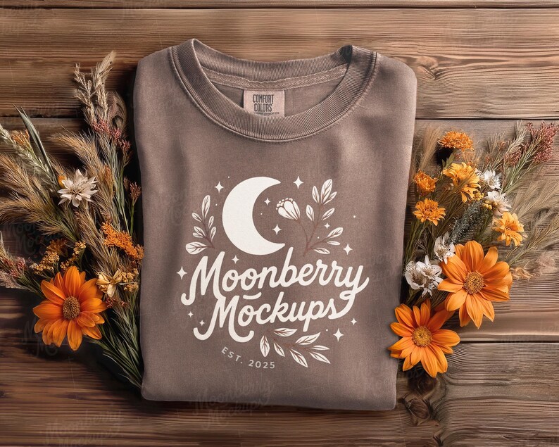 May include: A brown sweatshirt with a white crescent moon and floral design, with the text "Moonberry Mockups" and "EST. 2025". The sweatshirt is surrounded by orange flowers and dried plants on a wooden surface.