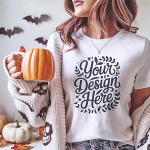 May include: White t-shirt with the text "Your Design Here" in a decorative font. The person is holding an orange pumpkin-shaped mug. Halloween-themed decor includes paper bats and pumpkins.