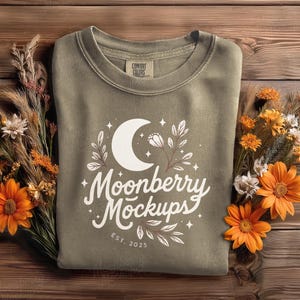 May include: An olive green sweatshirt with a white crescent moon and floral design, with the text "Moonberry Mockups EST. 2025". The sweatshirt is surrounded by orange flowers and dried plants on a wooden surface.