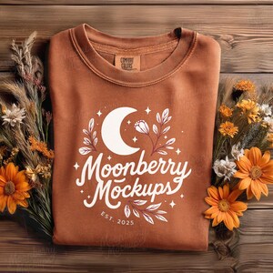 May include: A rust-colored sweatshirt with a white graphic featuring a crescent moon, floral elements, and the text "Moonberry Mockups EST. 2025." The sweatshirt is displayed on a wooden surface, adorned with dried flowers and orange blooms.