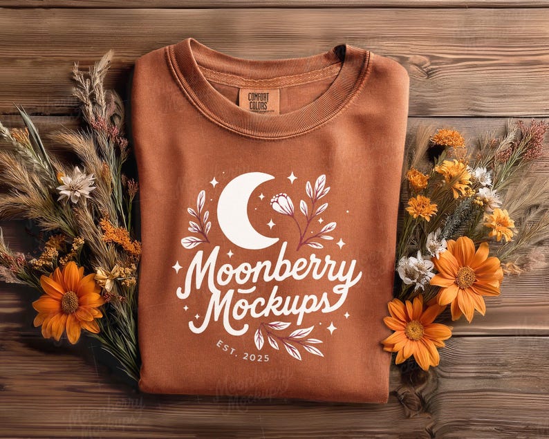 May include: A rust-colored sweatshirt with a white graphic featuring a crescent moon, floral elements, and the text "Moonberry Mockups EST. 2025." The sweatshirt is displayed on a wooden surface, adorned with dried flowers and orange blooms.