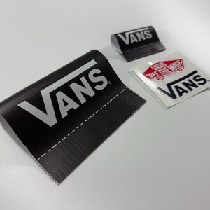 May include: A selection of Vans brand labels and stickers in black and white. The largest label is black with white text and a stitched edge. A smaller black label and a white sticker with red accents are also visible.
