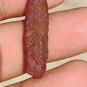 May include: A raw, elongated ruby crystal with a deep red hue and a textured, uneven surface. The gemstone has a columnar shape with natural facets and mineral inclusions, displaying a matte finish across its length and irregular edges.