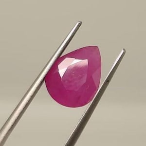 Könnte beinhalten: Ein facettierter, birnenförmiger Rubin-Edelstein, der von einer silbernen Pinzette gehalten wird. Der Rubin hat einen leuchtenden rosa-roten Farbton, mit mehreren Facetten, die das Licht einfangen. Die Pinzette ist so positioniert, dass die Form und Klarheit des Edelsteins gezeigt werden.