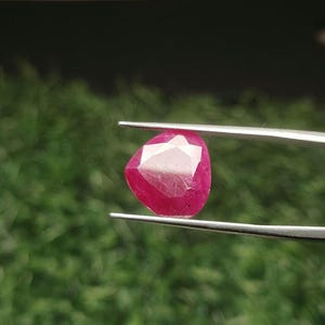 May include: A faceted, triangular-shaped ruby gemstone with a deep pinkish-red hue. The gemstone is held securely between the two thin, silver-colored metal tips of a pair of tweezers, showcasing its polished surface and geometric cut.