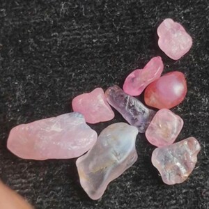 Puede incluir: Una colección de nueve piedras preciosas pequeñas, en bruto y translúcidas en varios tonos de rosa y lavanda pálido. Estas piedras tienen formas irregulares y pulidas con superficies lisas que captan la luz, mostrando variaciones naturales en la intensidad del color.