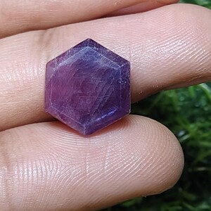 May include: A hexagonal, faceted gemstone with a deep purple hue. The crystal is held between fingers, showcasing its geometric shape and translucent quality. The gemstone's facets catch the light, creating a play of shadows and highlights.