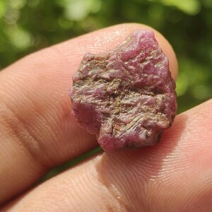 Op de afbeelding: Een ruwe, ongepolijste robijn edelsteen met een diepe magenta en roze tint. De steen heeft een ruwe, onregelmatige oppervlaktestructuur met zichtbare kristallijne structuren en aardse bruine insluitingen in de minerale vorm.