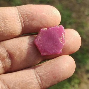 May include: A raw, vivid pink ruby gemstone. The ruby has a natural, irregular shape with a slightly textured surface. The gemstone is held between fingers, showcasing its colour and form. The background is blurred, suggesting an outdoor setting.