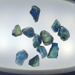 May include: A collection of eleven small, raw, unpolished gemstones with irregular shapes. The stones exhibit a translucent quality with a colour gradient ranging from deep blue to pale yellow-green, showing natural variations in their mineral structure.