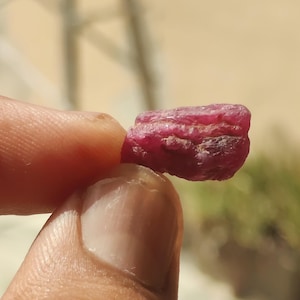 Puede incluir: Una piedra preciosa de rubí en bruto y sin pulir con un tono rojo magenta intenso. La piedra presenta una estructura cristalina natural e irregular con facetas, crestas y una superficie texturizada visibles. El rubí muestra variados tonos de rojo.