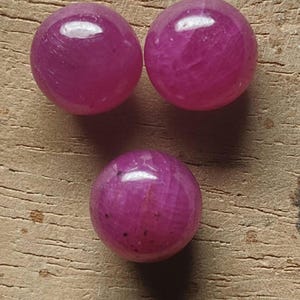 Op de afbeelding: Drie ronde, gepolijste kralen in een levendige magenta-roze tint. Elke kraal heeft een glad, glanzend oppervlak met subtiele interne kristallijne texturen en natuurlijke donkere insluitingen zichtbaar in het doorschijnende materiaal.