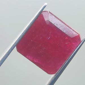 May include: A faceted, rectangular ruby gemstone held by tweezers. The ruby is a deep, saturated red colour with a slight sparkle. The gemstone's edges are precisely cut, showcasing its clarity and brilliance. The tweezers are silver.