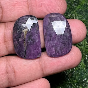 May include: Two polished, oval-shaped gemstones with a deep purple hue. Each stone features a faceted, geometric design on the top surface, creating a sparkling effect. The gemstones are held in a hand, showcasing their color and texture.