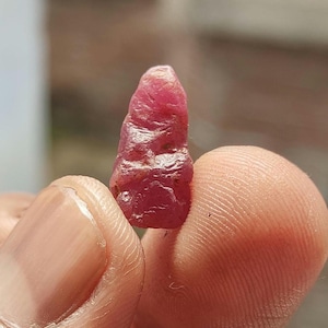 Puede incluir: Una piedra preciosa de rubí en bruto y sin pulir con un tono rojo rosado intenso. La piedra presenta una forma alargada e irregular con una superficie texturizada y facetada que muestra crestas y hendiduras naturales.