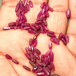Pode incluir: Uma coleção de pedras preciosas de rubi brutas e não cortadas. As pedras preciosas são de uma cor vermelha profunda e vibrante, com uma aparência ligeiramente translúcida. Os rubis são pequenos, alongados e de forma irregular, com um aspeto natural e não polido.
