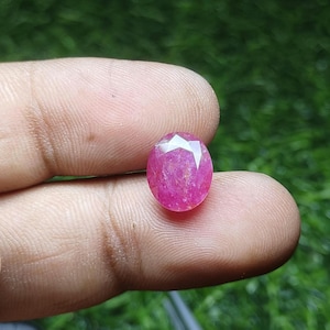 May include: An oval-shaped, faceted gemstone with a vibrant pinkish-red hue. The stone features a polished surface with multiple geometric facets that reflect light, showcasing a translucent interior with natural inclusions and a deep color saturation.