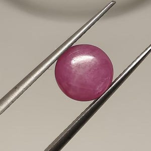 Op de afbeelding: Een ronde, gepolijste, roze edelsteen, vastgehouden door een zilveren pincet. De edelsteen heeft een glad oppervlak en een lichte glans, die het licht reflecteert. De achtergrond is een neutraal, lichtgrijs.