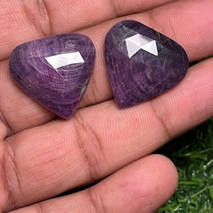 May include: Two heart-shaped, faceted gemstones with a deep purple hue. The gems have a polished surface with visible internal patterns and reflective facets. The stones are held in a hand, showcasing their color and shape.