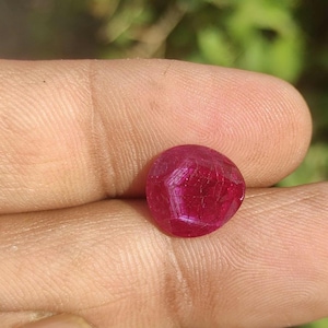 Puede incluir: Una gema de rubí redonda y facetada. La gema es de color rojo intenso con estructuras internas visibles. La piedra se sostiene en una mano, con un fondo verde borroso.