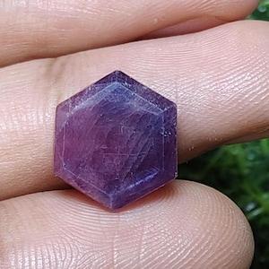 Puede incluir: Una gema hexagonal facetada de color púrpura intenso. La superficie de la gema refleja la luz, mostrando su estructura cristalina. La piedra se sostiene en una mano, resaltando su tamaño y forma. Adecuada para la fabricación de joyas o coleccionar.