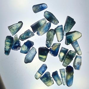 May include: A collection of loose, raw gemstones featuring a gradient of colours ranging from deep royal blue to pale yellow and translucent white. Each stone has an irregular, faceted shape with varying levels of clarity and internal inclusions.