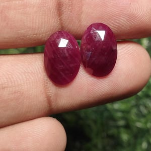 May include: Two oval-cut gemstones in a rich, deep red colour. The gems are held between fingers, highlighting their polished facets and internal sparkle. These are likely rubies, suitable for jewellery.