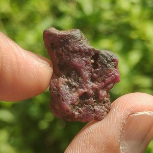Puede incluir: Una piedra preciosa de rubí en bruto y sin pulir con una coloración rojo intenso y granate oscuro. La piedra presenta una superficie irregular y texturizada con hoyos naturales, mostrando un acabado mate y un aspecto denso y opaco.