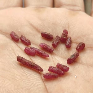 Peut inclure: Une collection de petits cristaux de rubis bruts rouge foncé, aux formes irrégulières et allongées avec des surfaces texturées. Ces pierres précieuses naturelles présentent une teinte cramoisie vive et un aspect brut non poli.