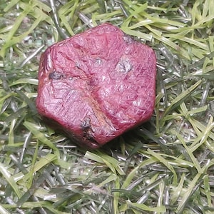 Peut inclure: Un cristal de rubis brut de forme hexagonale avec une couleur rouge profond et saturé et une surface texturée et inégale. La pierre précieuse présente des inclusions naturelles et un fini mat, révélant une forme géométrique à six côtés.