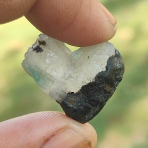 Pode incluir: Uma pedra preciosa bruta em forma de coração com uma mistura de cores claras, brancas e cinza escuro. A pedra é segurada entre dois dedos, mostrando sua textura natural e áspera. O fundo é verde desfocado.