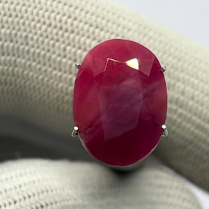 Può includere: Una grande gemma di rubino taglio ovale con una tonalità rosso intenso. La gemma è incastonata in una montatura in metallo argentato o bianco, con quattro griffe che fissano la pietra. Il rubino è il punto focale, mostrando le sue sfaccettature e la sua brillantezza.