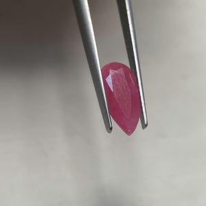 Könnte beinhalten: Ein birnenförmiger, facettierter rosa Edelstein, der von einer silberfarbenen Metallpinzette gehalten wird. Der Stein hat ein spitzes Ende und eine abgerundete Oberseite und zeigt eine durchscheinende Qualität mit sichtbaren Facetten.