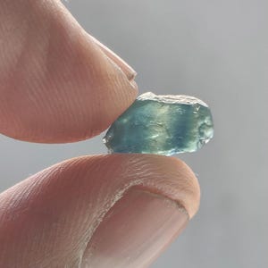 May include: A small, raw, translucent gemstone with a distinct colour gradient transitioning from a deep teal blue on one side to a lighter, pale green hue on the other. The stone has an irregular, multifaceted shape with a smooth, polished texture.