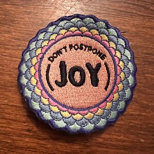 May include: A round, embroidered patch with a pink background and a colorful mandala border. The patch says "Don't Postpone (Joy)" in black text.