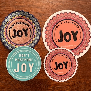 May include: Four round stickers with the text "Don't Postpone Joy" in different colors and designs. One sticker is embroidered with a pink background and a blue, yellow, and pink border. Another sticker has a pink background with a mandala design. The third sticker has a blue background with a white border. The fourth sticker has a pink background with a mandala design.