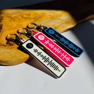 May include: Three keyrings in white, pink, and blue, each featuring a black and white barcode design. The keyrings have a rectangular shape with rounded edges and a silver keyring. The background is a wooden surface.