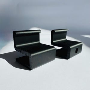 May include: Two black metal brackets with a unique, angular design. Each bracket features a curved top, a flat base, and a small hole. The brackets are positioned on a white surface, casting shadows.